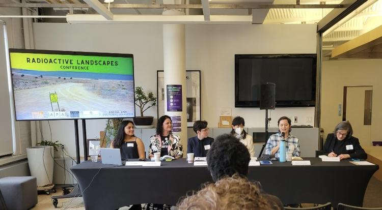 Ila Nako and Blue Carlsson sit with three other participants and a moderator at a long table. A screen behind them reads "Radioactive Landscapes."